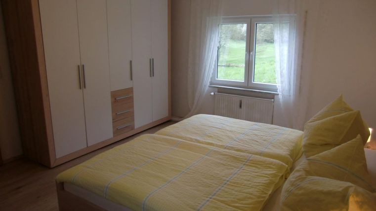 Une chambre confortable avec une grande fenêtre et un lit lumineux. L'armoire est moderne et le linge de lit est jaune.