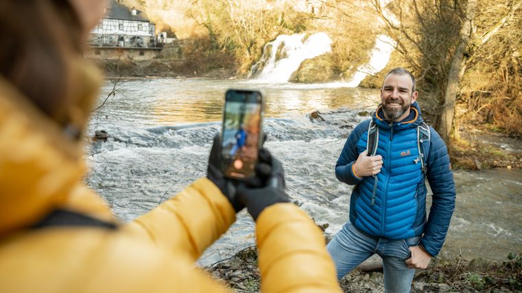 Ein Mann in blauer Jacke posiert lächelnd für ein Foto am Fluss, während eine andere Person ihn mit einem Smartphone fotografiert.