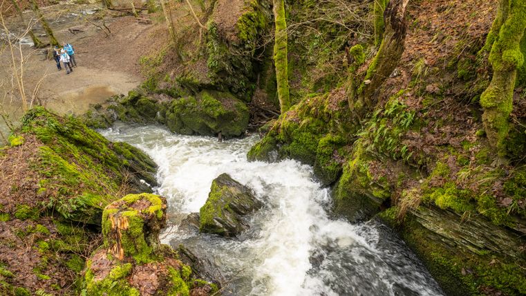 Schäumender Bachlauf in einer moosbewachsenen Schlucht entlang des Wanderwegs „Im Tal der wilden Endert“, Blick von oben auf Felsen, Wald und Wanderer im Hintergrund.