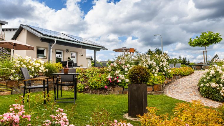 Ein gepflegter Garten mit bunten Blumen und einem gemütlichen Sitzbereich. Im Hintergrund steht ein modernes Haus unter einem bewölkten Himmel.