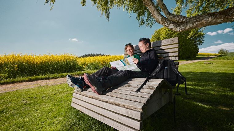Zwei Frauen entspannen sich auf einer Holzliege und schauen währenddessen in eine Wanderkarte.