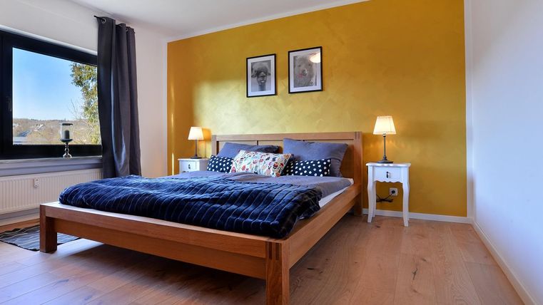 Een gezellige slaapkamer met een breed bed en decoratieve kussens. De muren zijn geschilderd in een warme gele tint en er zijn ramen met mooi uitzicht.