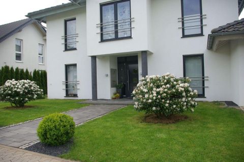 Ein modernes Haus mit einer hellen Fassade und großen Fenstern. Der gepflegte Vorgarten ist mit blühenden Sträuchern und einem kleinen Buchsbaum angelegt.
