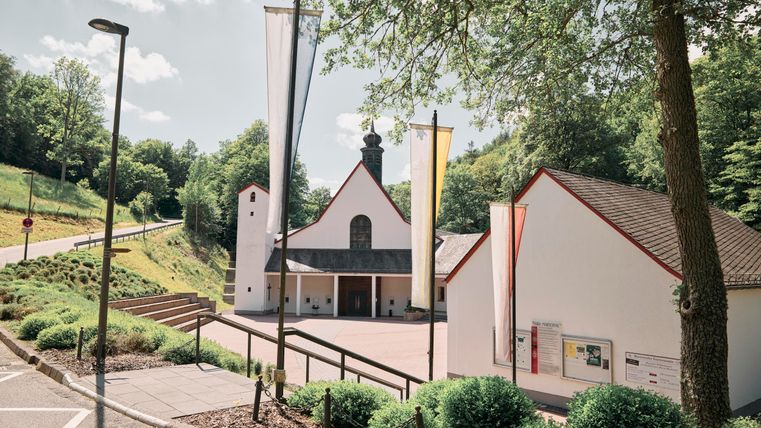 Ein modernes Gebäude mit einem markanten Eingang und hellen Fahnen. Umgeben von grünen Büschen und Bäumen, unter einem klaren Himmel.