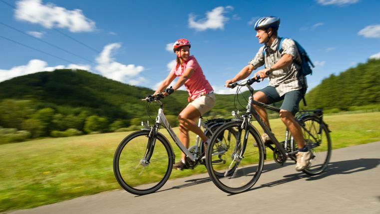 Zwei Radfahrer fahren auf einer asphaltierten Straße. Im Hintergrund grüne Wiesen und bewaldete Hügel.
