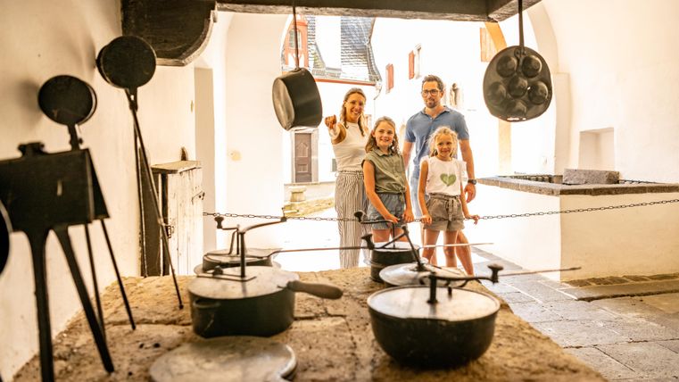 Eine Familie steht in einer historischen Küche mit verschiedenen Töpfen und Pfannen. Im Hintergrund sind weitere Räume sichtbar, die den alten Charme des Ortes ausstrahlen.