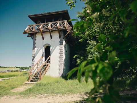 Ein historisches Gebäude mit einem Holzbalustrade und einer Eingangstreppe. Es steht umgeben von Grün und Bäumen in einer ländlichen Umgebung.