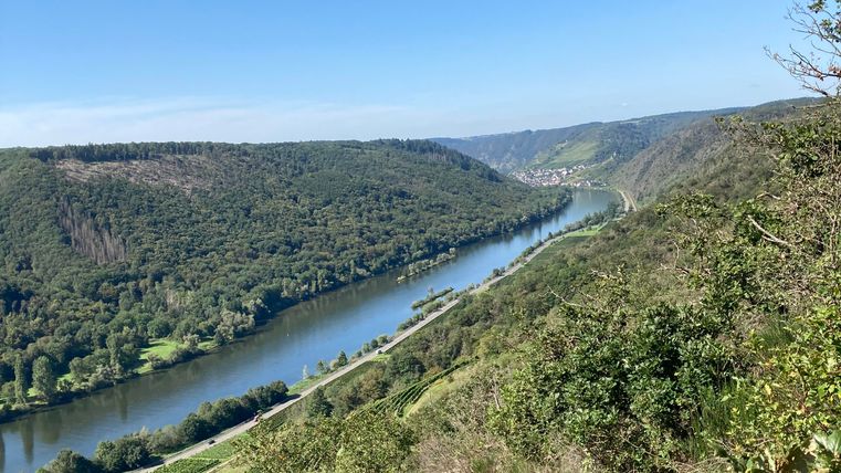 Eine malerische Landschaft mit einem Fluss, umgeben von grünen Hügeln. Der Himmel ist klar und blau.