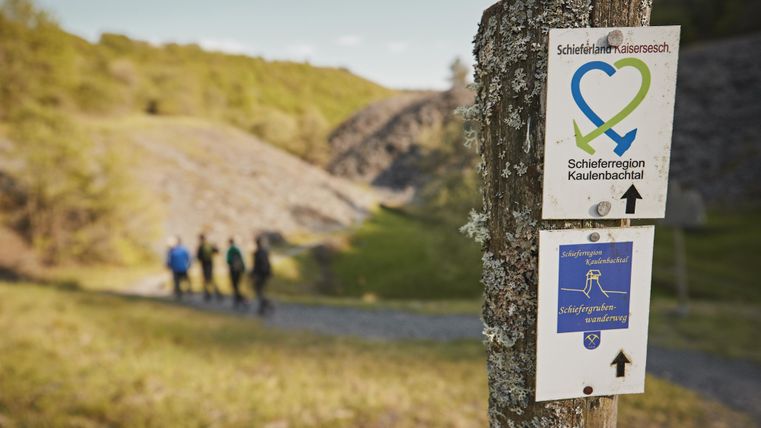 Ein Wegweiser auf einem Wanderweg zeigt die Richtung an. Im Hintergrund sind wandernde Personen in einer naturnahen Umgebung sichtbar.