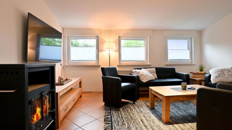 A cozy living room with a modern television, comfortable sofas, and a table. The large windows provide plenty of daylight.