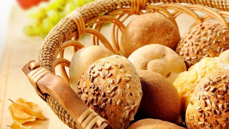 Ein Korb mit verschiedenen Brötchen liegt auf einem Tisch. Im Hintergrund sind ein Apfel und Trauben zu sehen.