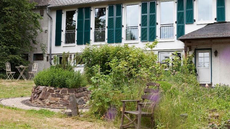 Une maison confortable avec des volets verts et un jardin bien entretenu. Au premier plan se trouve un banc en bois entouré de plantes.