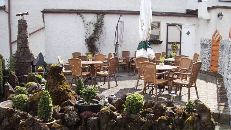 Een gezellige terras met houten tafels en stoelen. Omringd door een verzorgde tuin met kleine planten en een decoratief steenelement.
