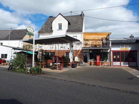 Ein kleines Dorf mit einem gemütlichen Gasthaus und einer Outdoor-Sitzgelegenheit. Im Vordergrund gibt es einen Tisch mit Stühlen und eine alte Zapfsäule.