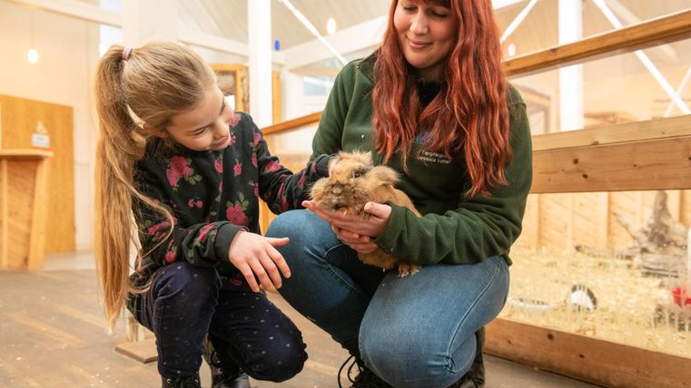 Ein Mädchen und eine Frau halten ein Kaninchen in einem Tiergarten. Beide lächeln und zeigen Freude beim Interagieren mit dem Tier.