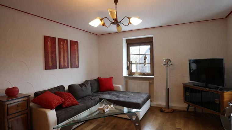 Ein gemütliches Wohnzimmer mit einer dunklen Couch, roten Kissen und einem Glastisch. An der Wand hängen dekorative Bilder, und ein Fernseher steht neben einem Fenster.