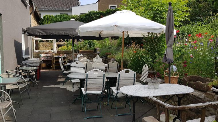 Ein gemütlicher Außenbereich mit Tischen und Stühlen. Umbrellas sorgen für Schatten, umgeben von üppigem Grün und Blumen.