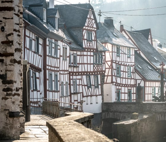 Fachwerkhäuser in Monreal, Deutschland, mit roten Balken und weißen Wänden. Eine Brücke führt über einen kleinen Fluss, im Hintergrund bewaldete Hügel., © Eifel Tourismus GmbH, D. Ketz