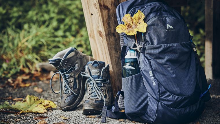 Ein Rucksack steht neben einem Paar Wanderschuhe auf einem Gehweg. Am Rucksack ist ein gelbes Blatt befestigt, umgeben von grünem Hintergrund.