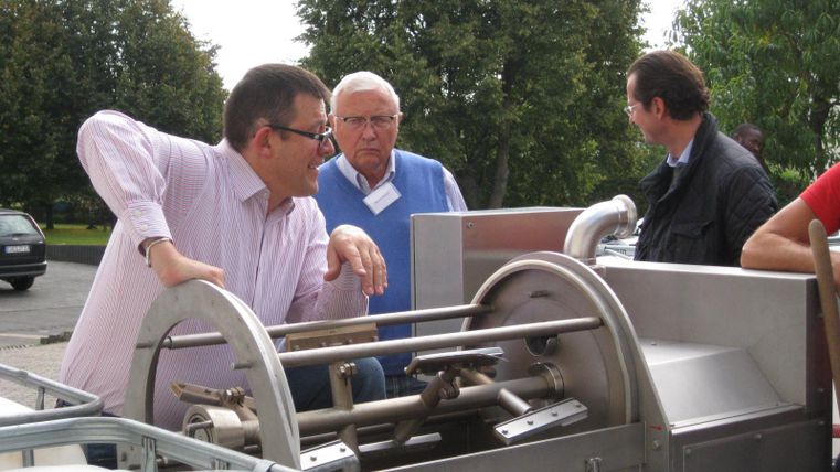 Een groep mannen bespreekt een machine in de buitenlucht. Op de achtergrond zijn bomen en enkele voertuigen te zien.