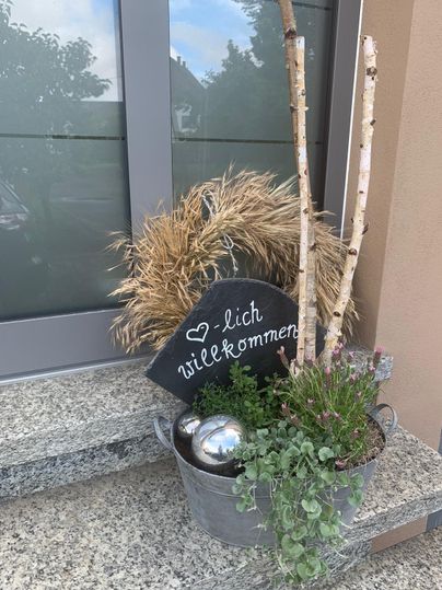 Eine dekorative Pflanzenschale mit einem Willkommensschild. Die Schale enthält Gräser, Zweige und schöne Metallkugeln.