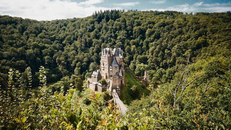 Die Burg Eltz mit ihren markanten Türmen und Fachwerkbauten liegt eingebettet in ein dicht bewaldetes Tal, umgeben von Sommergrün und blauem Himmel.