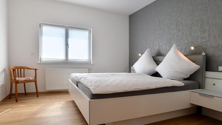 Ein modernes Schlafzimmer mit einem großen Bett und zwei Kissen. Neben dem Bett steht ein Holzstuhl und ein Fenster sorgt für natürliches Licht.