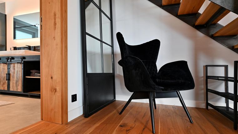 Ein moderner Sessel steht in einem stilvollen Raum mit Holzfußboden. Im Hintergrund ist eine Treppe und eine Glaswand zu sehen.