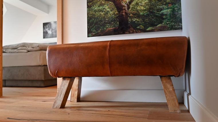 Ein stilvoller, brauner Lederhocker mit Holzbeinen steht an einer Wand. Im Hintergrund ist ein Bild eines Baumes zu sehen.