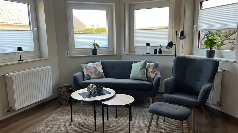 Ein modernes Wohnzimmer mit einem grauen Sofa und einem Sessel. Es gibt große Fenster, einen Teppich und dekorative Tischchen.