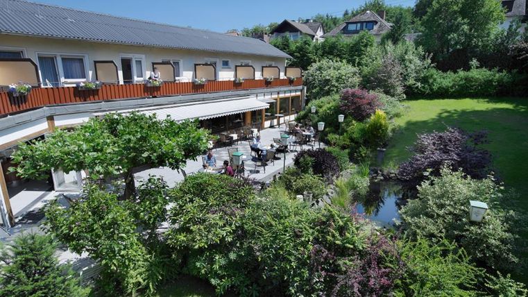 Ein ruhiger Garten mit vielen Pflanzen und Bäumen umgibt ein gemütliches Gebäude. Auf der Terrasse stehen Tische und Stühle, ideal für entspannte Stunden im Freien.