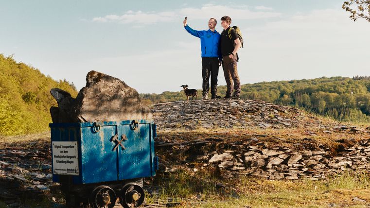 Two hikers with a dog take a selfie on a slate field in the Eifel. On the left in the foreground is a blue wagon with slate rock, behind it forests and green hills under a blue sky.