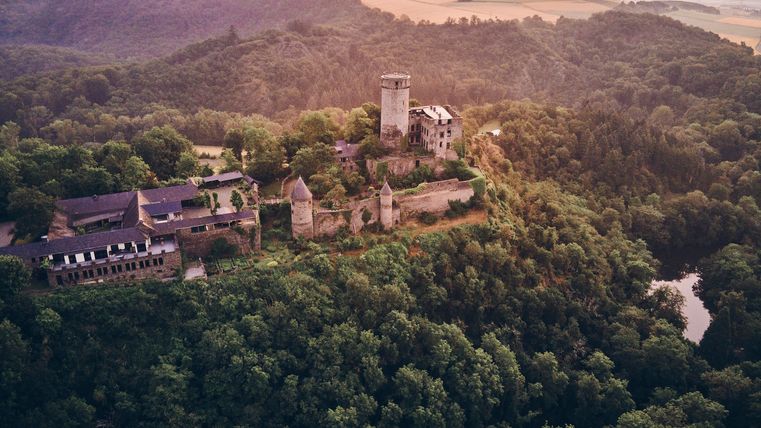 Luftaufnahme einer mittelalterlichen Burg auf einem bewaldeten Hügel bei Sonnenaufgang; die Burg mit markantem Turm und dicken Mauern erhebt sich über ein grünes Tal, im Hintergrund sanfte Hügel und ein pastellfarbener Himmel.