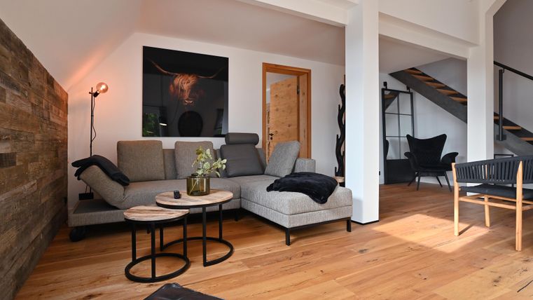 Ein modernes Wohnzimmer mit einem grauen Sofa, einem Couchtisch und Holzfußboden. Die Wände sind hell, und es gibt eine Treppe im Hintergrund.