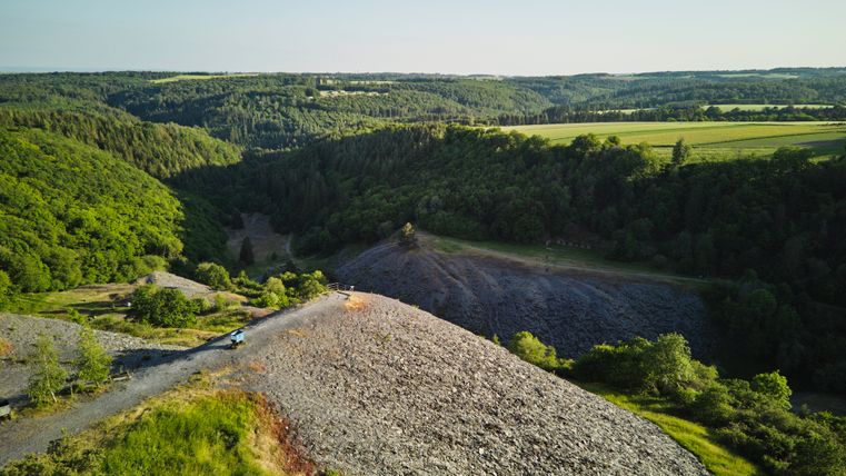 Blick über das Kaulenbachtal in der Eifel: Im Vordergrund eine grüne Wiese, daneben ein graues Schieferfeld. Dahinter erstrecken sich bewaldete Hügel und Täler bis zum Horizont unter blauem Himmel.
