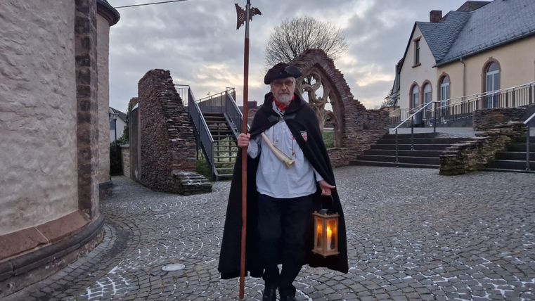Ein Mann in traditioneller Kleidung steht auf einem gepflasterten Platz. Er hält eine Laterne und einen Stock in der Hand, umgeben von historischer Architektur.