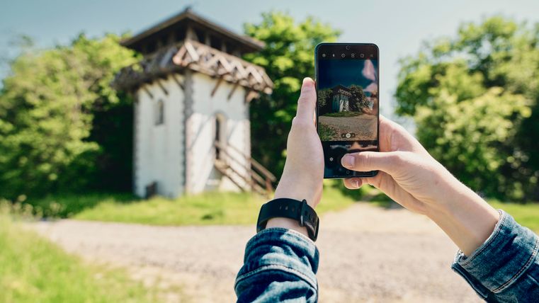 Eine Person fotografiert ein historisches Gebäude mit einem Smartphone. Im Hintergrund sind Bäume und ein klarer Himmel zu sehen.